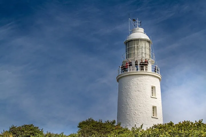 Turistas de pie en el balcón del Cape Bruny Lighthouse blanco contra un cielo azul.